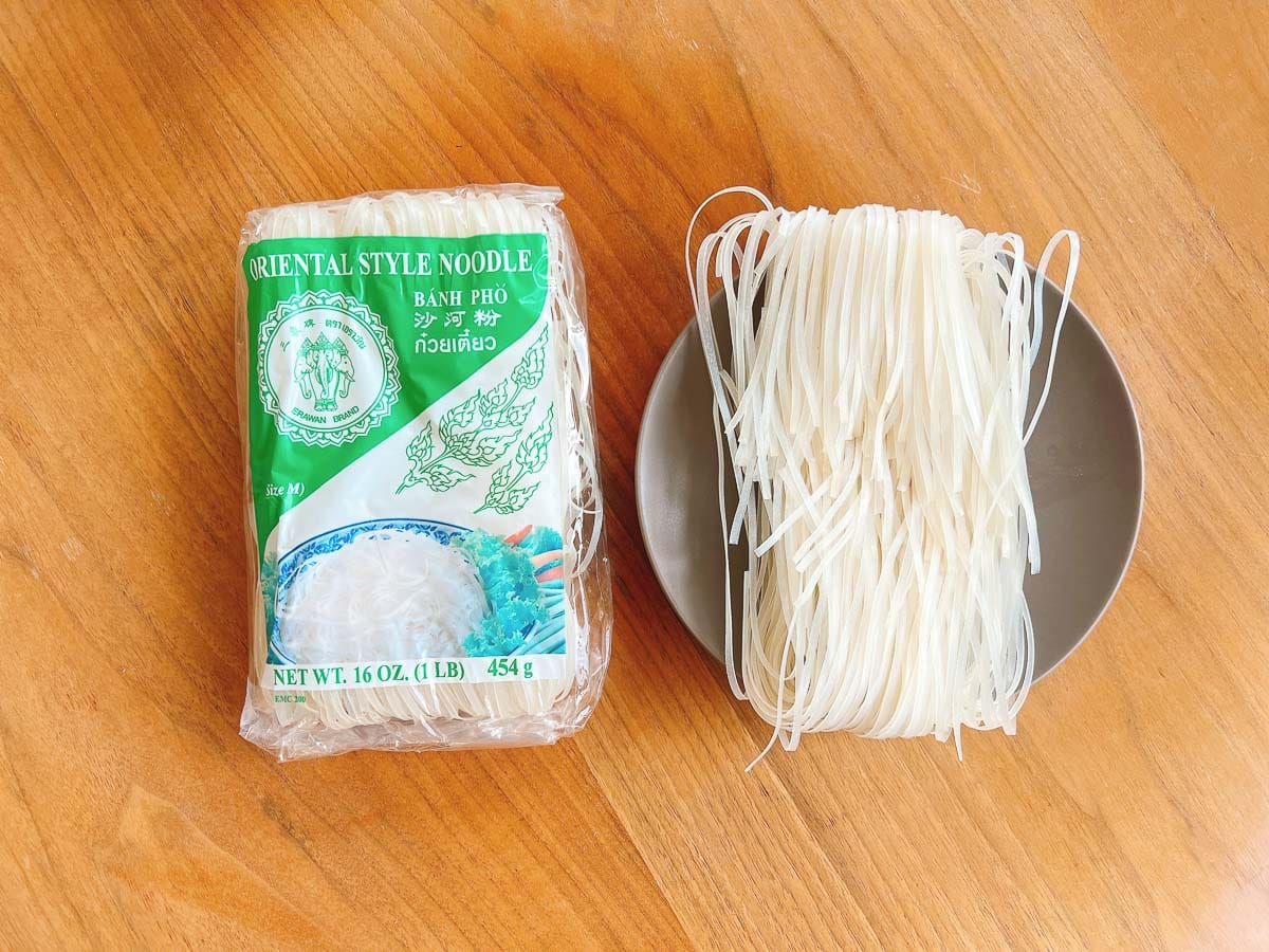 A package of oriental style pho rice noodles sits next to a bowl containing uncooked rice noodles, all placed on a wooden surface—perfect for pairing with rich Turkey pho bone broth.