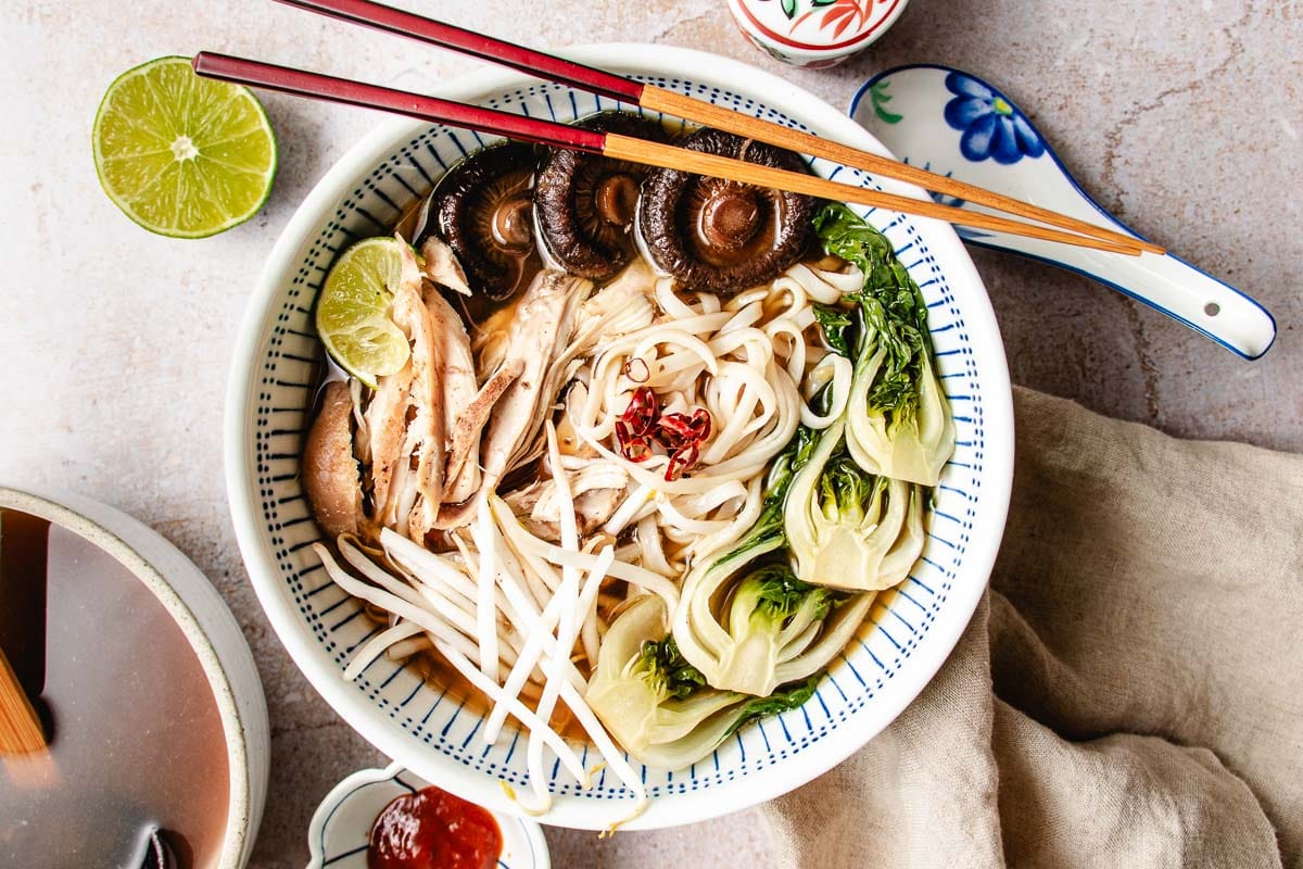 A bowl of noodle soup with mushrooms, shredded turkey, bok choy, bean sprouts, lime wedges, and red chili in a rich turkey pho bone broth, served with chopsticks and a ceramic spoon on the side.