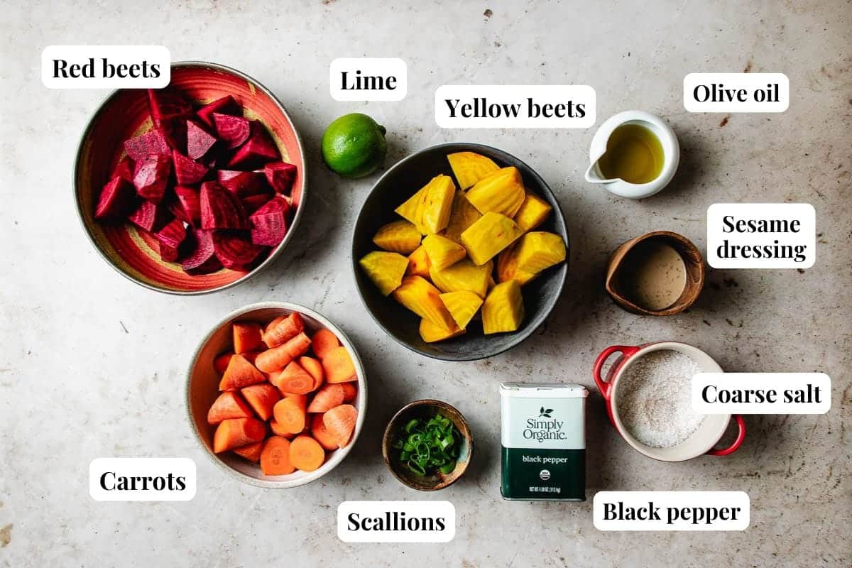 Bowls of roasted beets and carrots, scallions, a whole lime, olive oil, sesame dressing, coarse salt, and black pepper are arranged on a white surface with labels for each ingredient.