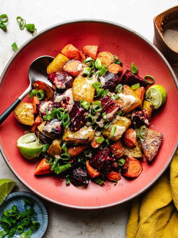 A red plate filled with roasted beets and carrots, and green onions garnish with drizzled with creamy dressing, garnished with lime wedges and served with a spoon on the side.