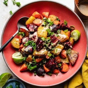 A red plate filled with roasted beets and carrots, and topped with chopped green onions and a creamy sauce. Lime wedges and a spoon are beside the vibrant vegetables.