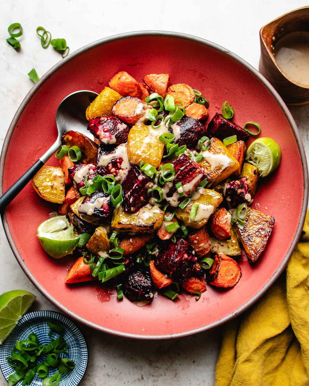 A red plate filled with roasted beets and carrots, and green onions garnish with drizzled with creamy dressing, garnished with lime wedges and served with a spoon on the side.