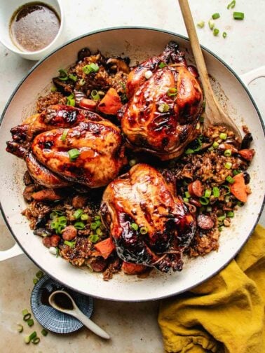 A white baking dish with three roasted Cornish hens, glazed and nestled on a bed of rice stuffing with sausage, and vegetables, garnished with green onions. A wooden spoon rests inside, with a cup of sauce and a yellow napkin nearby.