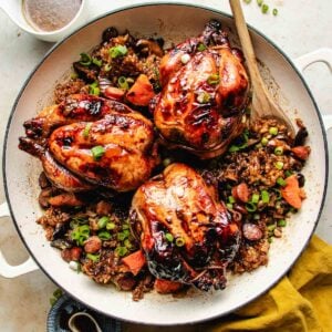 Three roasted Cornish hens sit atop a hearty sticky rice stuffing with sausage, carrots, and green onions in a large white skillet. A wooden spoon rests on the side, and a small bowl of sauce is nearby.