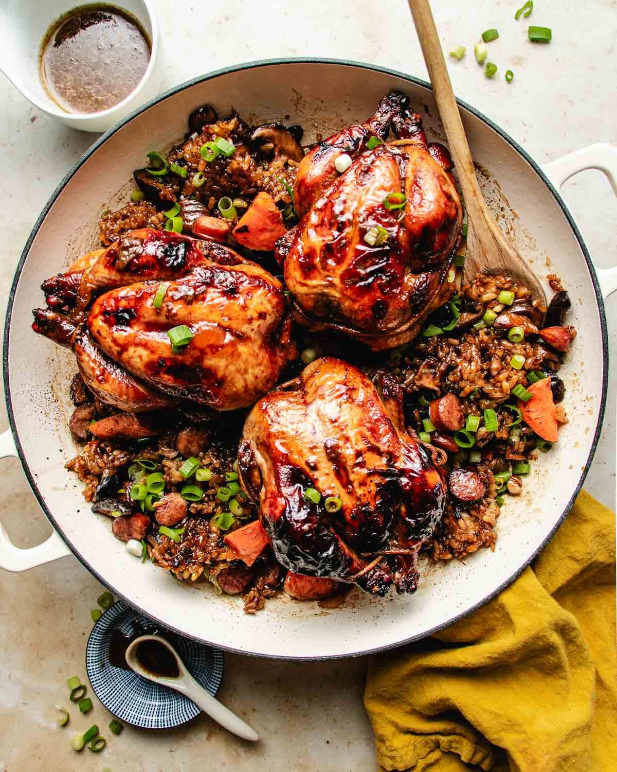A white baking dish with three roasted Cornish hens, glazed and nestled on a bed of rice stuffing with sausage, and vegetables, garnished with green onions. A wooden spoon rests inside, with a cup of sauce and a yellow napkin nearby.