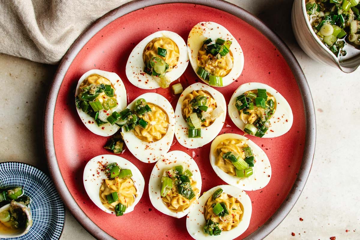 A plate of Asian deviled eggs topped with chopped green onions , scallion oil, and herbs, arranged on a red dish. A small bowl of extra topping is nearby, and part of a blue plate is visible in the corner.