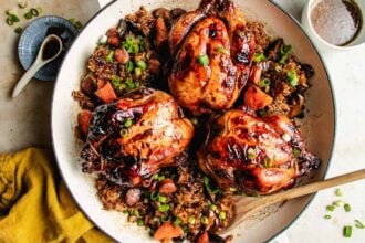 Three roasted Cornish hens, glazed to perfection, sit atop a bed of seasoned sticky rice with sausage slices, vegetables, and chopped green onions in a large white dish. A wooden spoon rests nearby, ready to serve this savory feast.