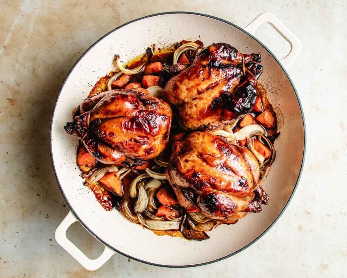 Three roasted Cornish hens in a white pan with caramelized onions and chunks of carrots, all browned, glistening, and bursting with flavor, set on a light surface.
