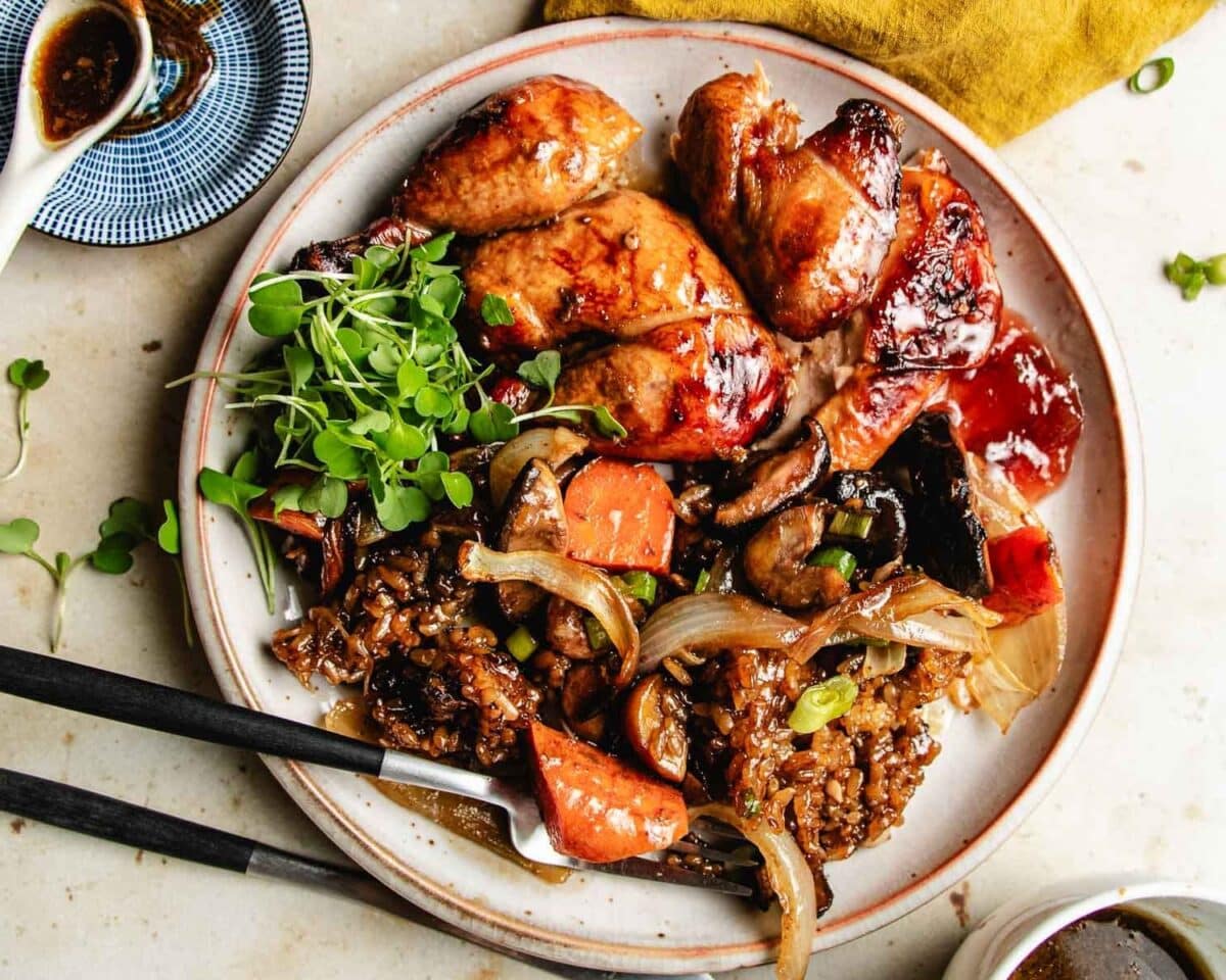 A plate of glazed roasted Cornish hens, with stuffing on the plate - vegetables, rice, and microgreens, with chopsticks resting on the side. A small bowl of sauce is partially visible in the background.