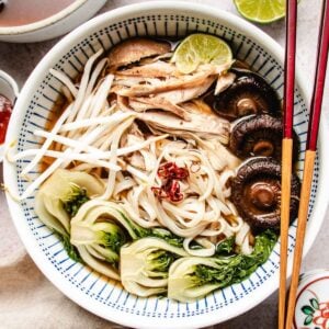 A bowl of noodle soup in rich turkey pho bone broth, topped with shredded turkey, mushrooms, bok choy, bean sprouts, sliced red chili, and a lime wedge, served with chopsticks on the side.