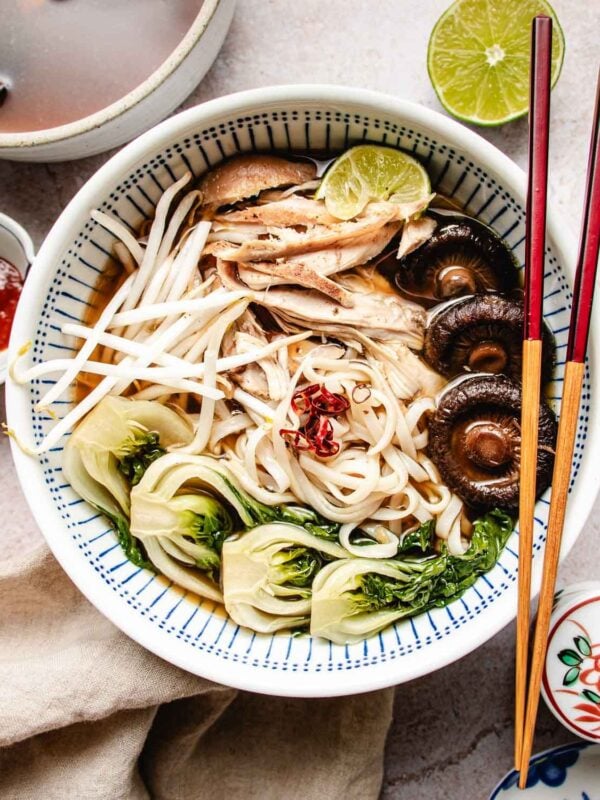 A bowl of noodle soup topped with bok choy, mushrooms, bean sprouts, shredded turkey, a lime wedge, and red chili slices in savory turkey pho bone broth, with chopsticks resting on the bowl.