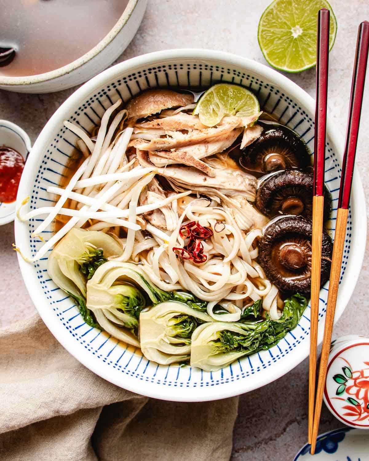 A bowl of noodle soup topped with bok choy, mushrooms, bean sprouts, shredded turkey, a lime wedge, and red chili slices in savory turkey pho bone broth, with chopsticks resting on the bowl.