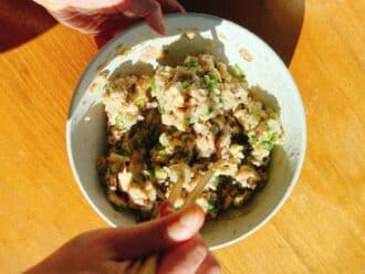 Hands mixing a mixture of vegetables, meat, and herbs for Asian wonton meatballs in a white bowl using chopsticks, with sunlight casting shadows on a wooden table.