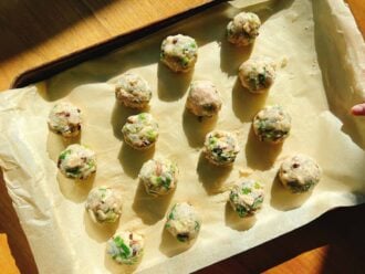 A baking tray lined with parchment paper holds rows of uncooked Asian wonton meatballs mixed with chopped herbs and vegetables. Sunlight casts gentle shadows across the wooden surface and tray.