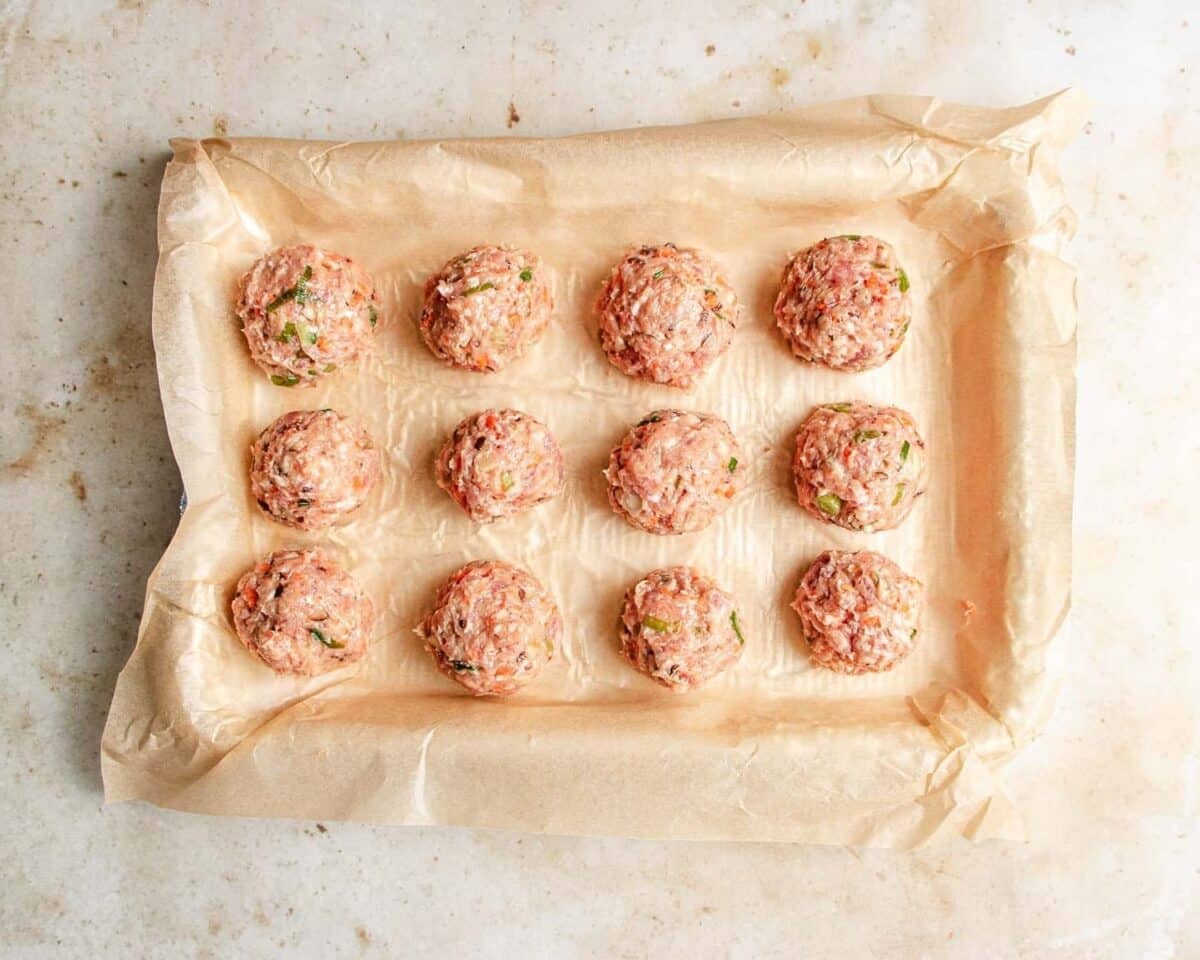 Twelve raw meatballs with green herbs, inspired by deconstructed wonton soup, are evenly spaced on a parchment-lined baking tray, ready to be cooked. The tray sits on a light-colored surface.