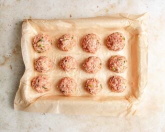Twelve raw meatballs with green herbs, inspired by deconstructed wonton soup, are evenly spaced on a parchment-lined baking tray, ready to be cooked. The tray sits on a light-colored surface.