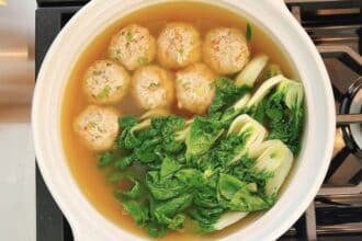 A deconstructed wonton soup features meatballs, chopped bok choy, and leafy greens gently simmering in a clear broth on the stove.
