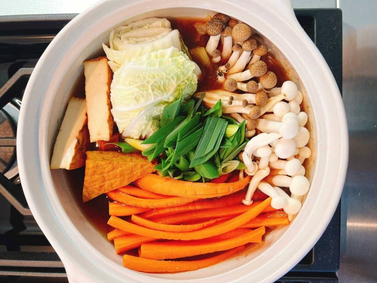 A pot on a stove filled with neatly arranged vegetables, tofu, and chicken meatball hot pot ingredients like napa cabbage, carrots, mushrooms, green onions, and fried tofu, all ready to be cooked.