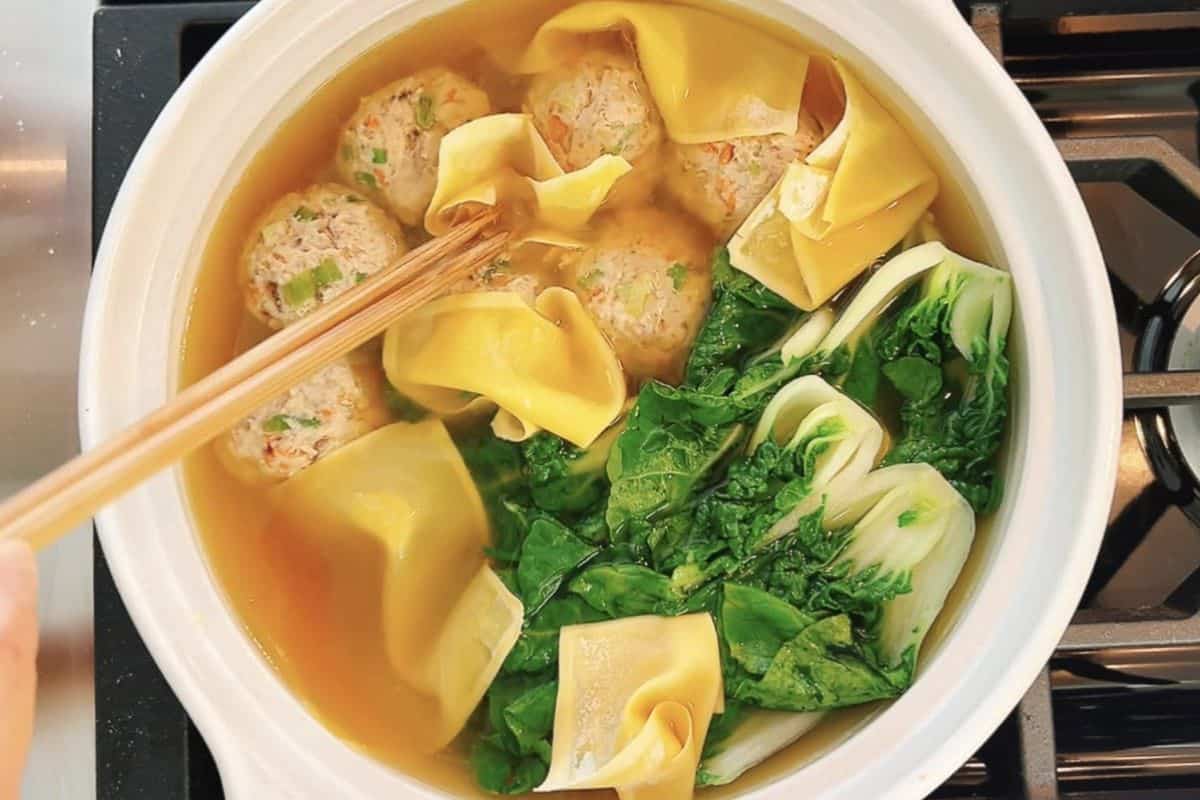 A pot of deconstructed wonton soup simmers on the stove, with meatballs, yellow dumpling wrappers, and leafy green vegetables being stirred gently with chopsticks.