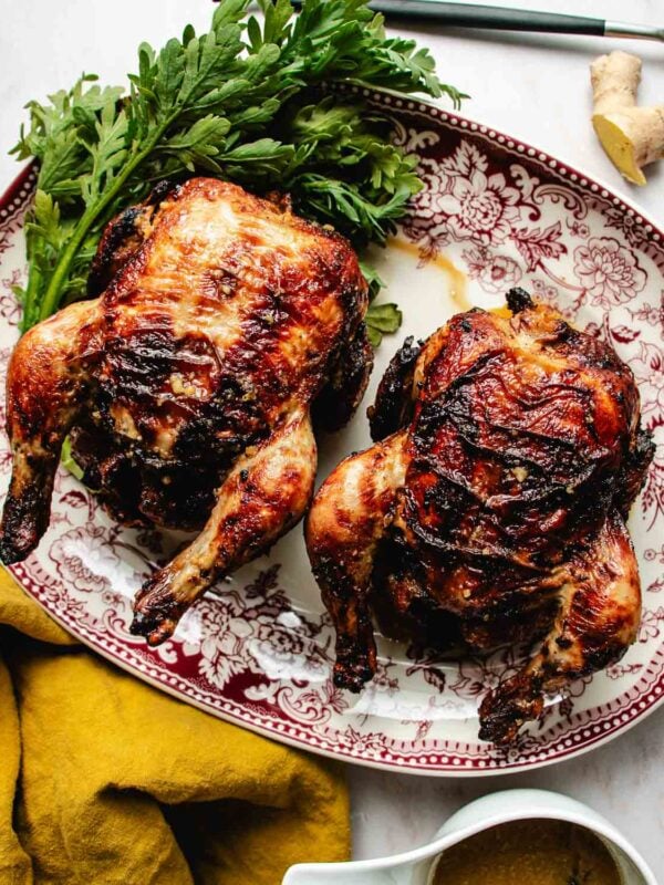 Two roasted air fryer Cornish hens with browned, crispy skin are served on a decorative red and white platter, garnished with fresh green herbs. A yellow napkin and a piece of ginger are nearby.