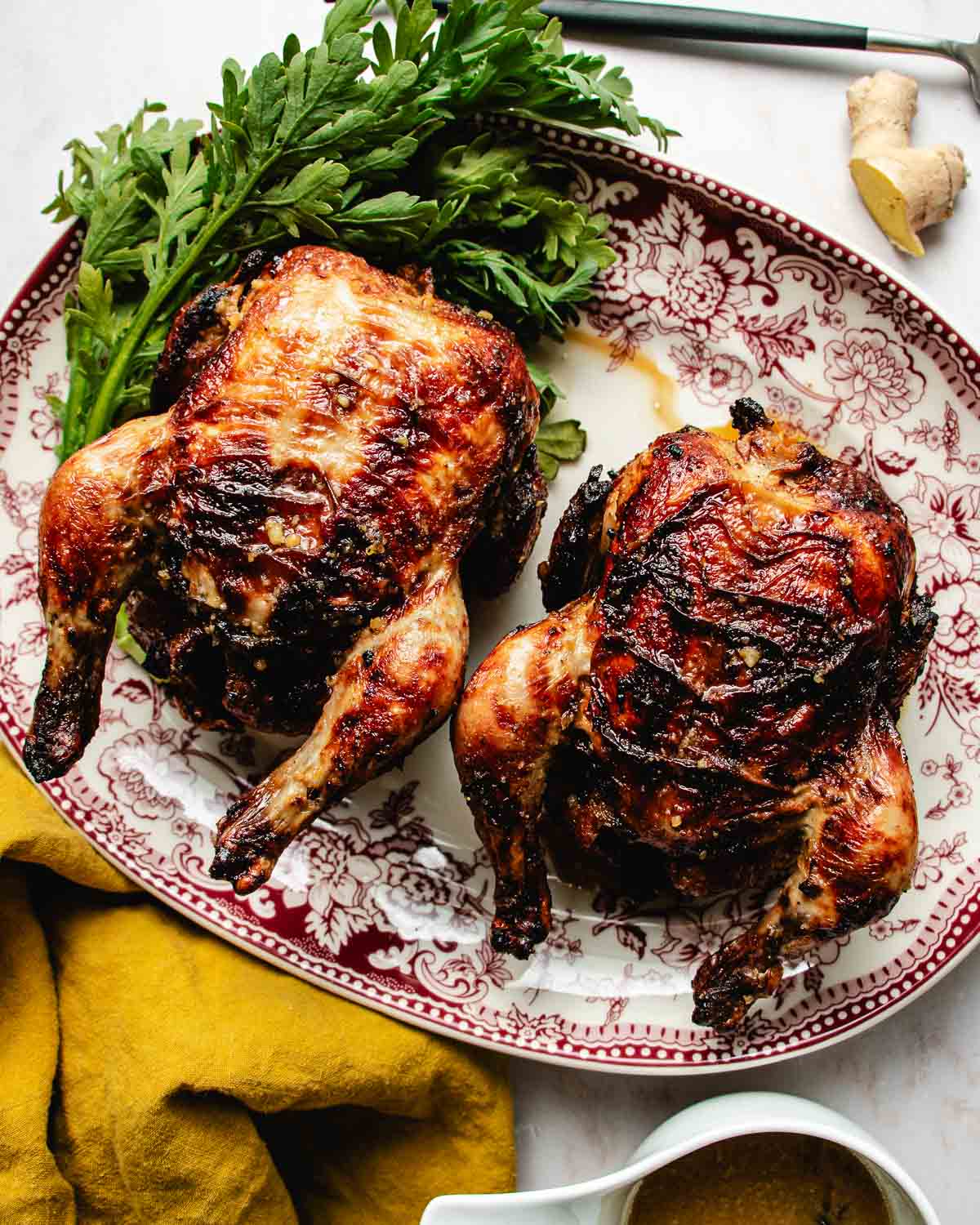 Two roasted air fryer Cornish hens with browned, crispy skin are served on a decorative red and white platter, garnished with fresh green herbs. A yellow napkin and a piece of ginger are nearby.
