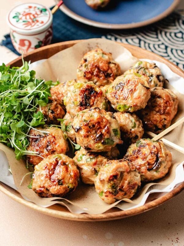 A wooden bowl lined with parchment holds several golden-brown Asian wonton meatballs garnished with green herbs. Fresh microgreens sit on the side, and skewers are visible among the savory meatballs.