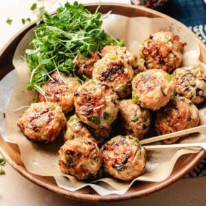 A wooden bowl lined with parchment paper holds several browned Asian wonton meatballs with herbs, accompanied by a small bunch of fresh microgreens. A skewer rests on the side of the bowl.