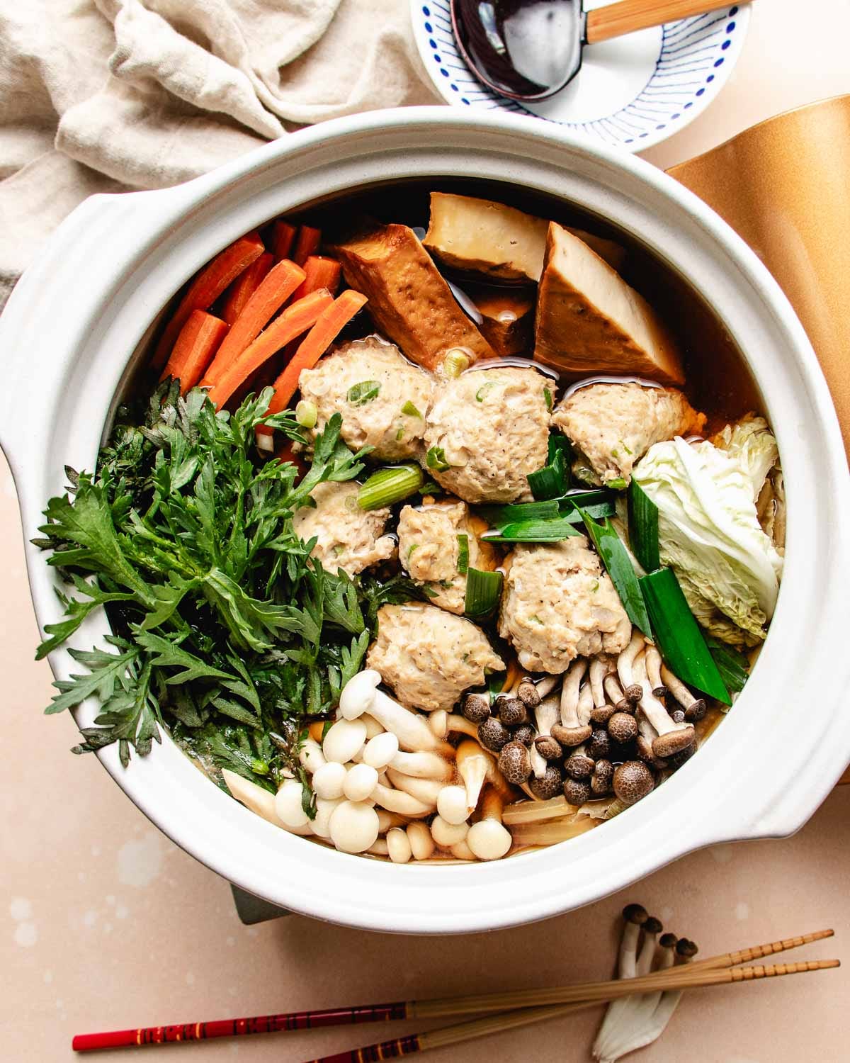 A pot of chicken meatball hot pot filled with assorted vegetables, mushrooms, leafy greens, tofu, carrot sticks, and chicken meatballs, all arranged neatly in a flavorful broth. Chopsticks and a spoon are beside the pot.