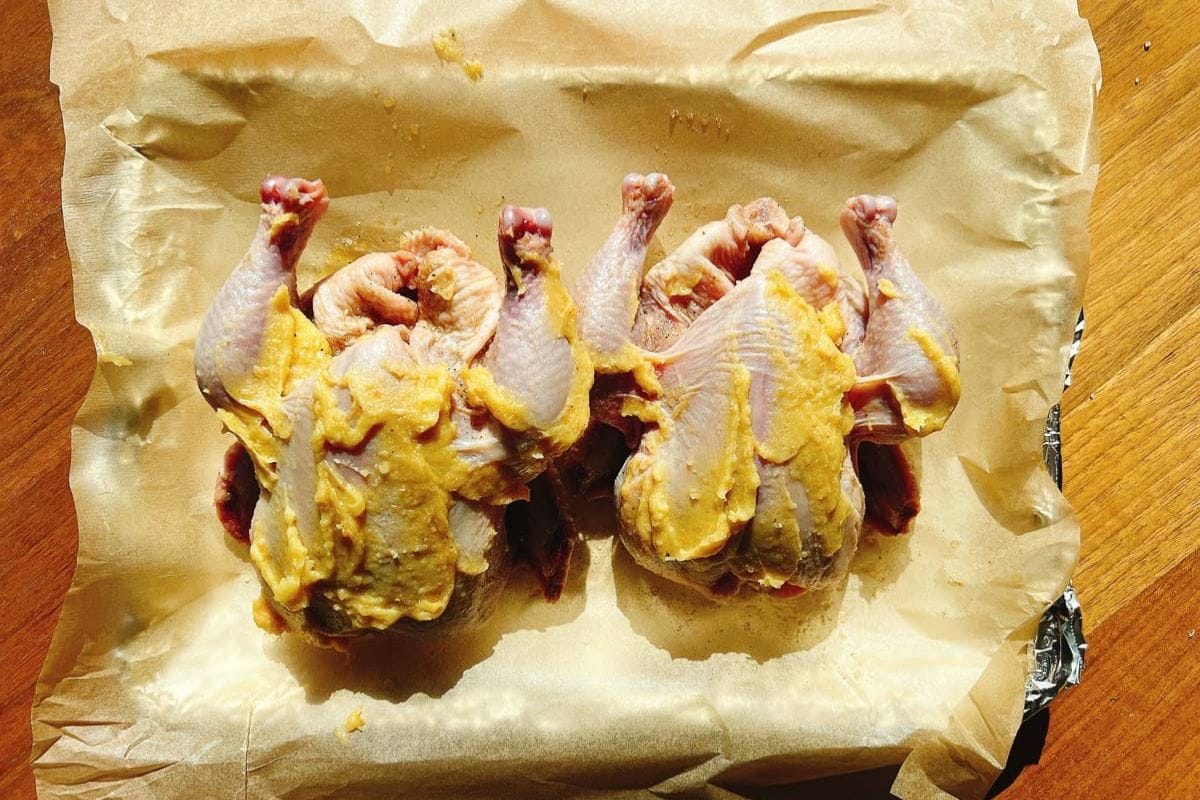 Two raw, whole Cornish hens covered in a yellow colored ghee butter mixed with miso paste sit side by side on parchment paper atop a baking tray, ready for cooking. The scene is brightly lit by natural sunlight—perfect inspiration for your next air fryer Cornish hens recipe.