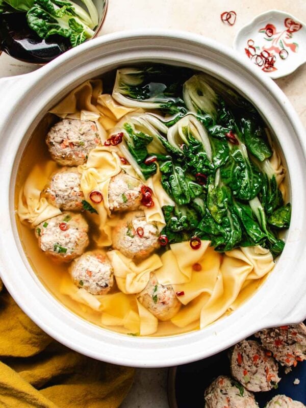 A bowl of deconstructed wonton soup features wonton wrappers, meatballs, bok choy, and sliced red chili peppers in a light broth, all beautifully presented in a white pot on a table.