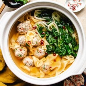 A deconstructed wonton soup featuring clear broth, bok choy, wonton wrappers, and meatballs, garnished with sliced red chilies and served elegantly in a white pot.