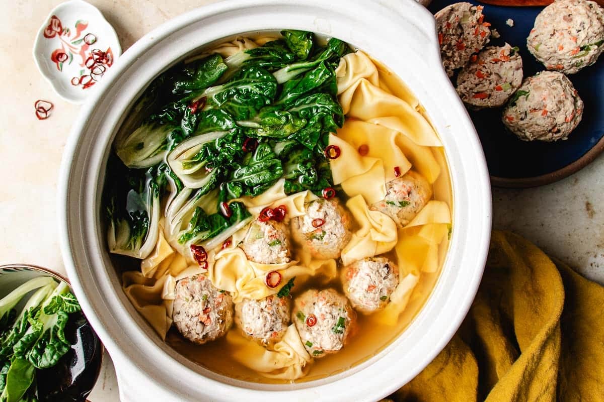A bowl of deconstructed wonton soup with wonton sheets, baby bok choy, and several wonton meatballs in a flavorful broth, garnished with sliced red chili peppers. Extra raw meatballs rest on a plate nearby, accompanied by fresh greens and a yellow cloth.