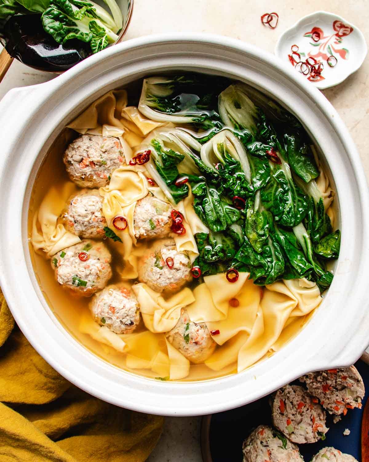 A bowl of deconstructed wonton soup features wonton wrappers, meatballs, bok choy, and sliced red chili peppers in a light broth, all beautifully presented in a white pot on a table.