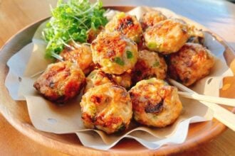 A wooden bowl lined with parchment paper holds several golden-brown Asian wonton meatballs with chopped green herbs, accompanied by fresh green sprouts and wooden picks.