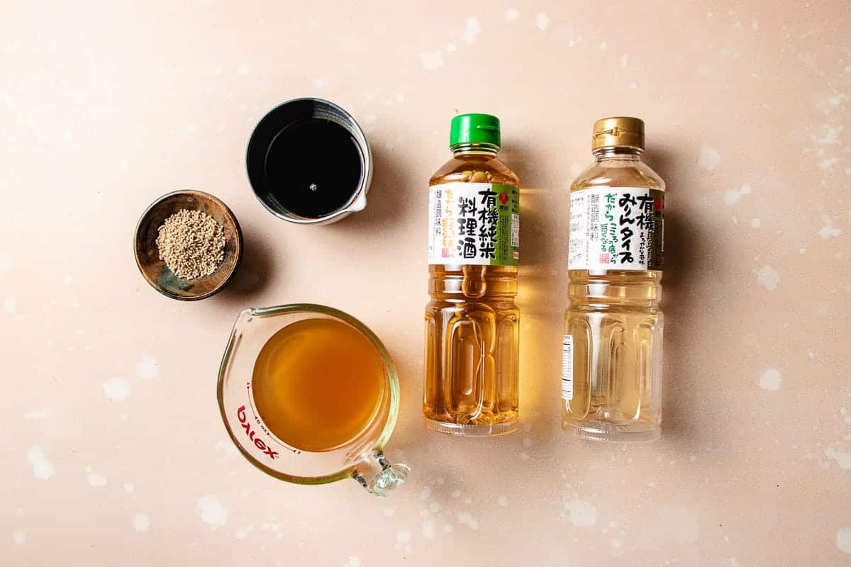 Overhead view of two bottles with Japanese mirin and sake, a measuring cup of soy sauce, a small bowl of dashi powder, and a metal measuring cup with dark liquid—ingredients perfect for preparing chicken meatball hot pot—on a beige surface with white spots.