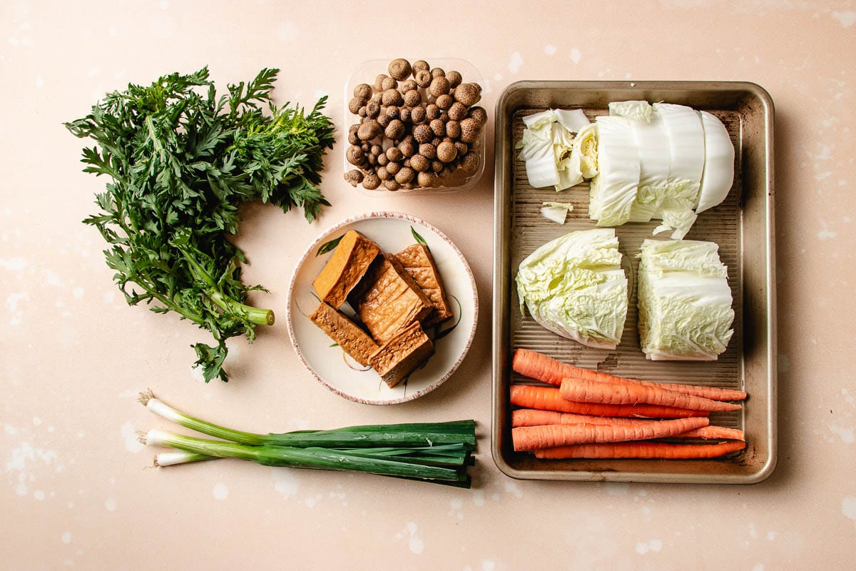 A top-down view of fresh vegetables for chicken meatball hot pot on a light surface: leafy greens, a bowl of tofu, mushrooms, green onions, carrot sticks, and napa cabbage wedges on a baking tray.