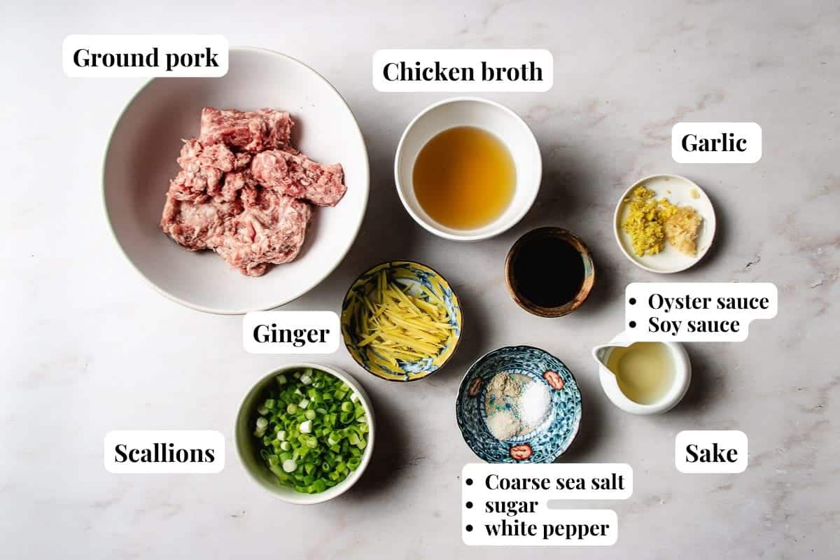 A flat lay of labeled ingredients in bowls for a recipe: ground pork, chicken broth, minced garlic, sliced ginger, scallions, oyster sauce, soy sauce, sake, coarse sea salt, sugar, and white pepper—perfect for homemade xiao long bao soup dumplings.