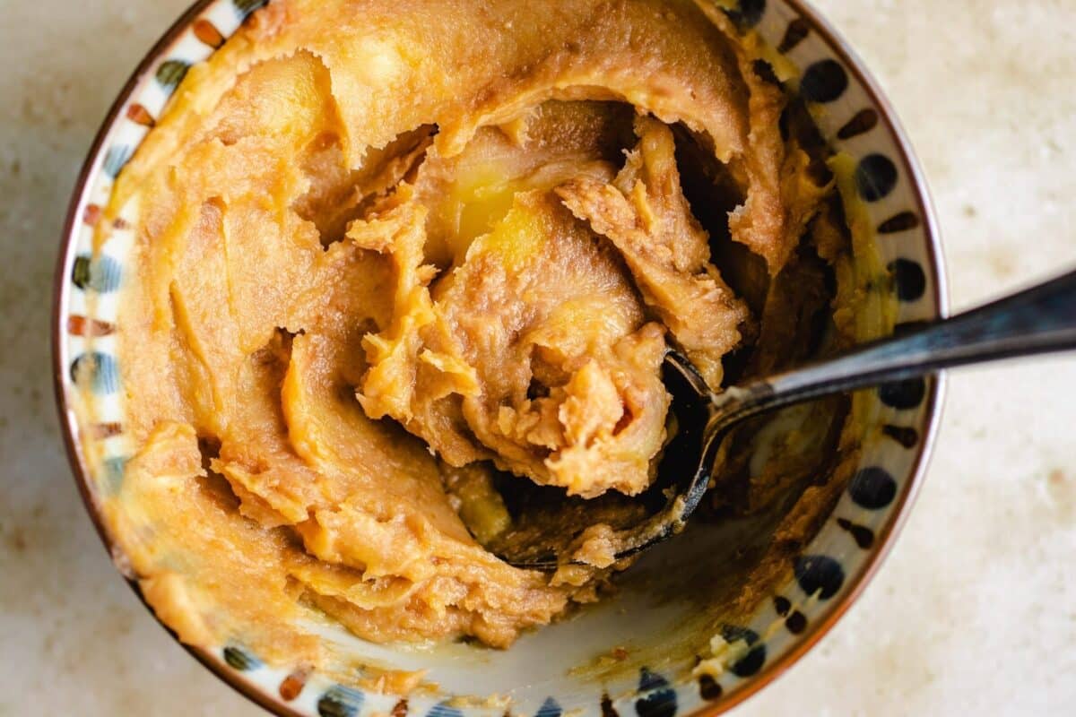 A close-up of a bowl filled with creamy, amber-colored miso butter paste, with a spoon partially submerged and some paste clinging to it—perfect for glazing air fryer Cornish hens. The bowl has a decorative rim and sits on a light surface.