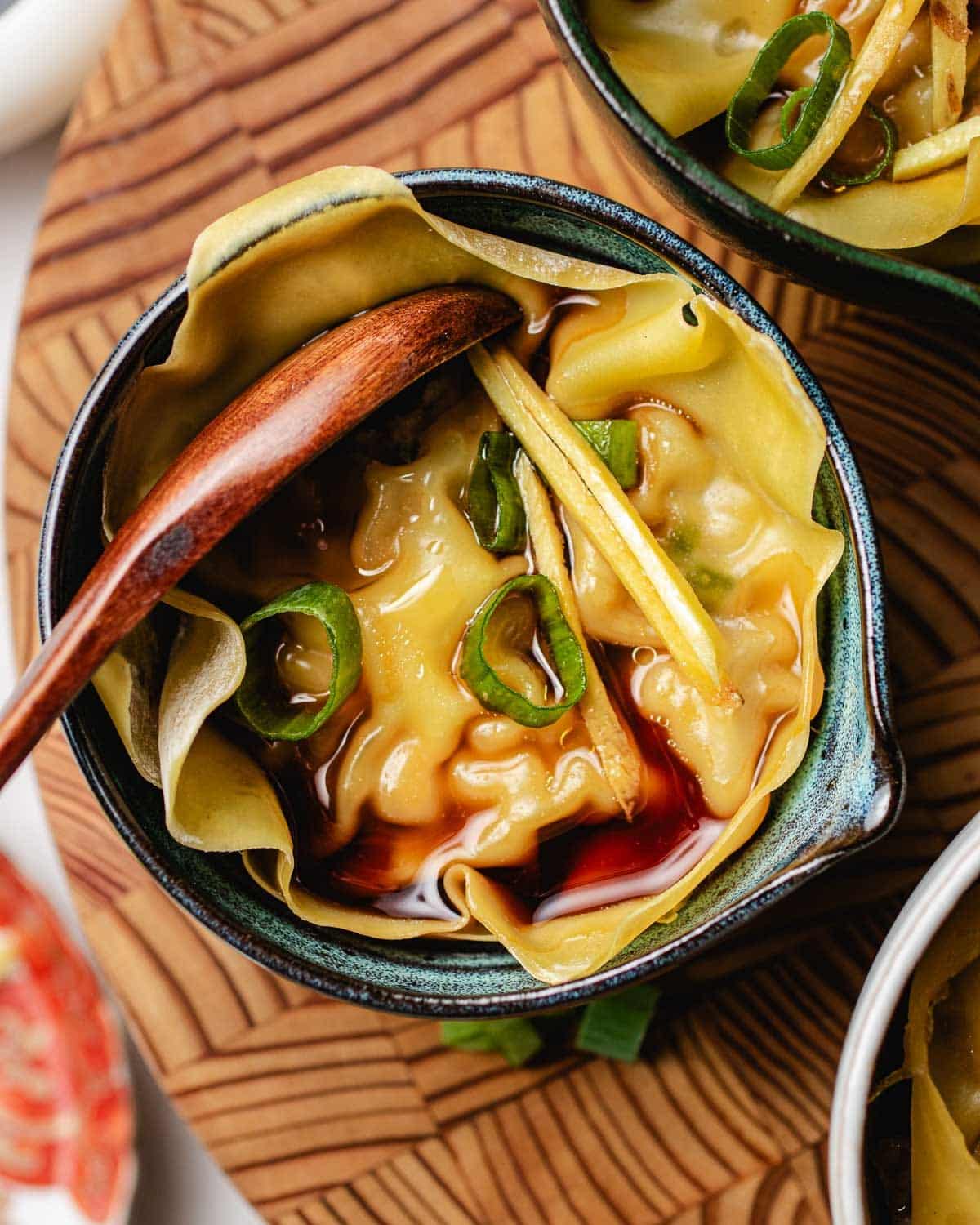 A bowl of soup dumplings lasagna topped with sliced green onions and ginger, with a wooden spoon resting inside. The xiaolongbao is covered in broth and a drizzle of dark sauce.