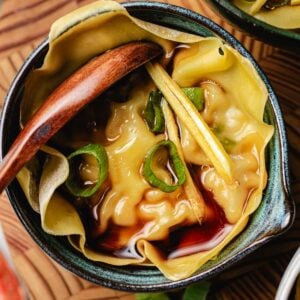 A close-up of a soup dumpling xiaolongbao in broth, garnished with sliced green onions and thin ginger strips, served in a small ceramic bowl with a wooden spoon.