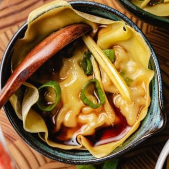 A close-up of a soup dumpling xiaolongbao in broth, garnished with sliced green onions and thin ginger strips, served in a small ceramic bowl with a wooden spoon.