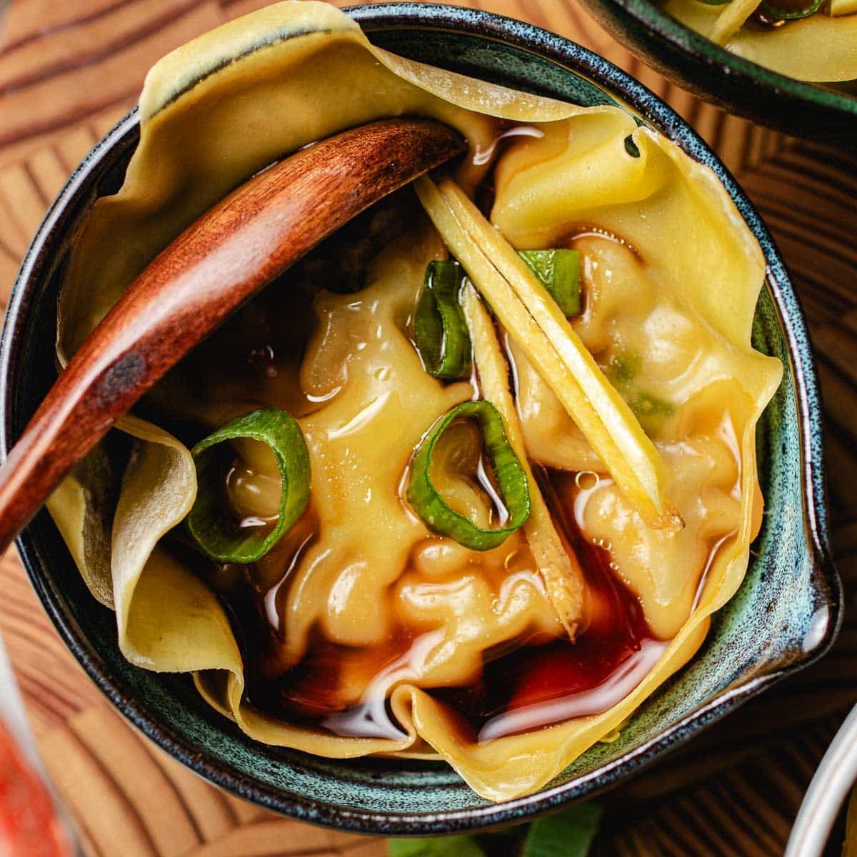 A close-up of a soup dumpling xiaolongbao in broth, garnished with sliced green onions and thin ginger strips, served in a small ceramic bowl with a wooden spoon.