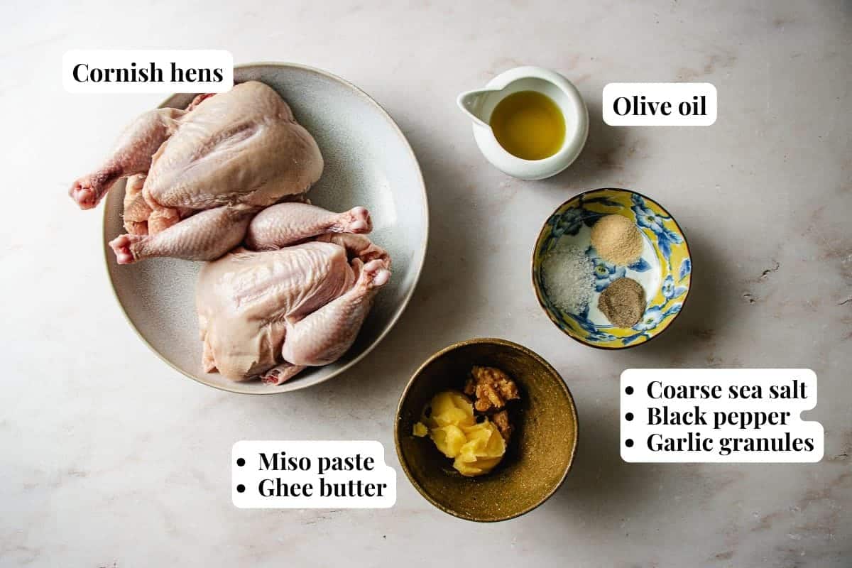 Two raw Cornish hens on a plate, ready to become delicious air fryer Cornish hens. Bowls of olive oil, miso paste, ghee butter, coarse sea salt, black pepper, and garlic granules surround them—each ingredient clearly labeled.