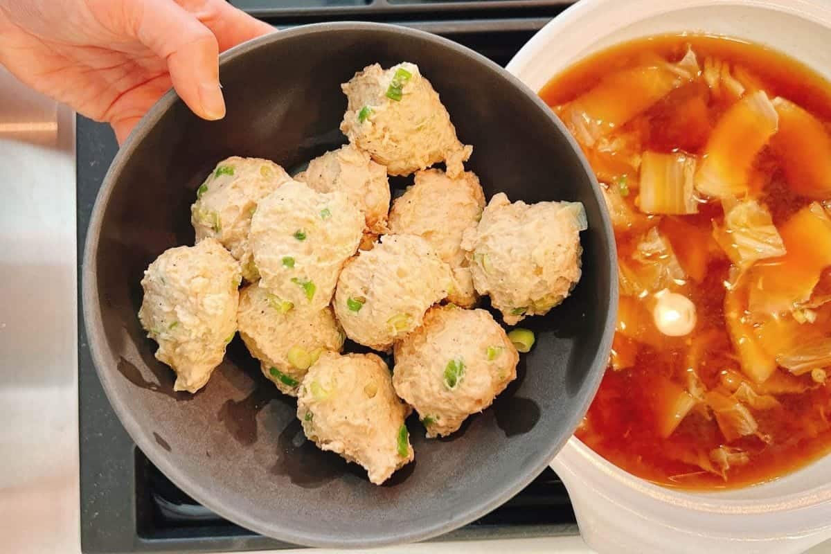 A hand holds a black bowl filled with simmered homemade chicken meatball hot pot, garnished with chopped green onions, next to a pot of soup containing cabbage in a rich brown broth.