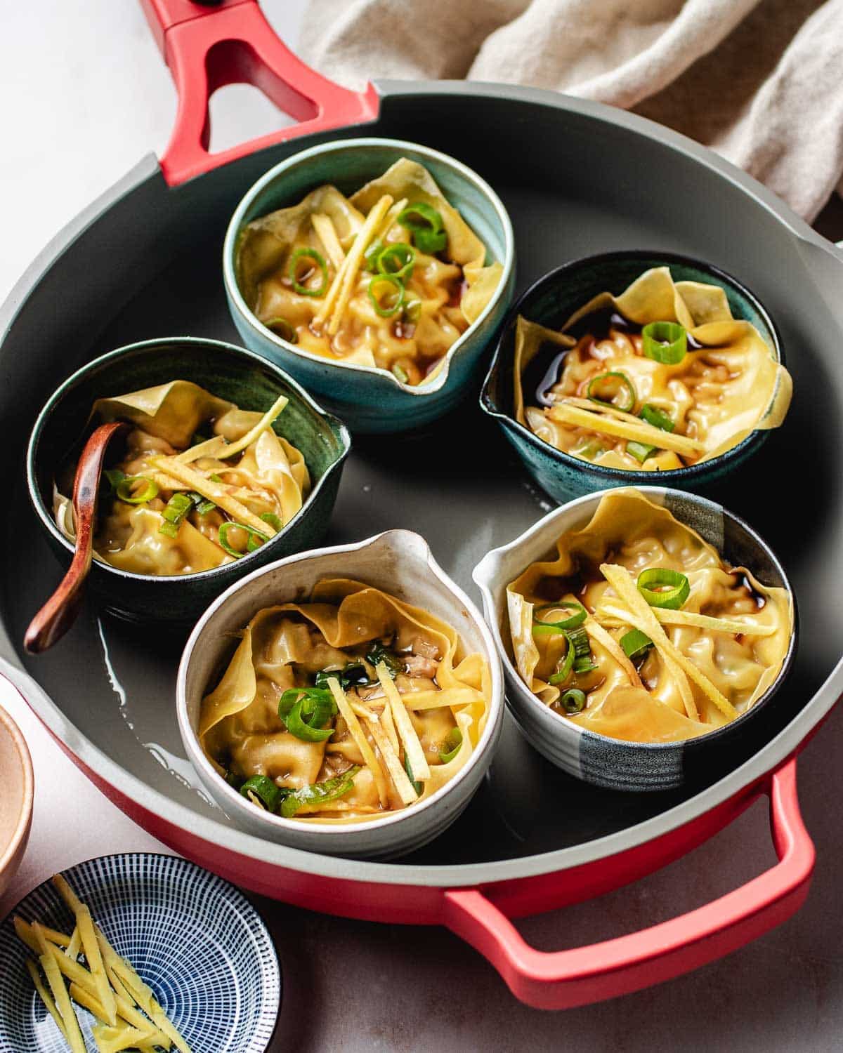 Five bowls of soup dumplings in small cups, garnished with sliced green onions and ginger, are arranged inside a large red pan. A wooden spoon rests in one bowl, and a beige napkin is in the background. These xiaolongbao promise rich, comforting flavors.