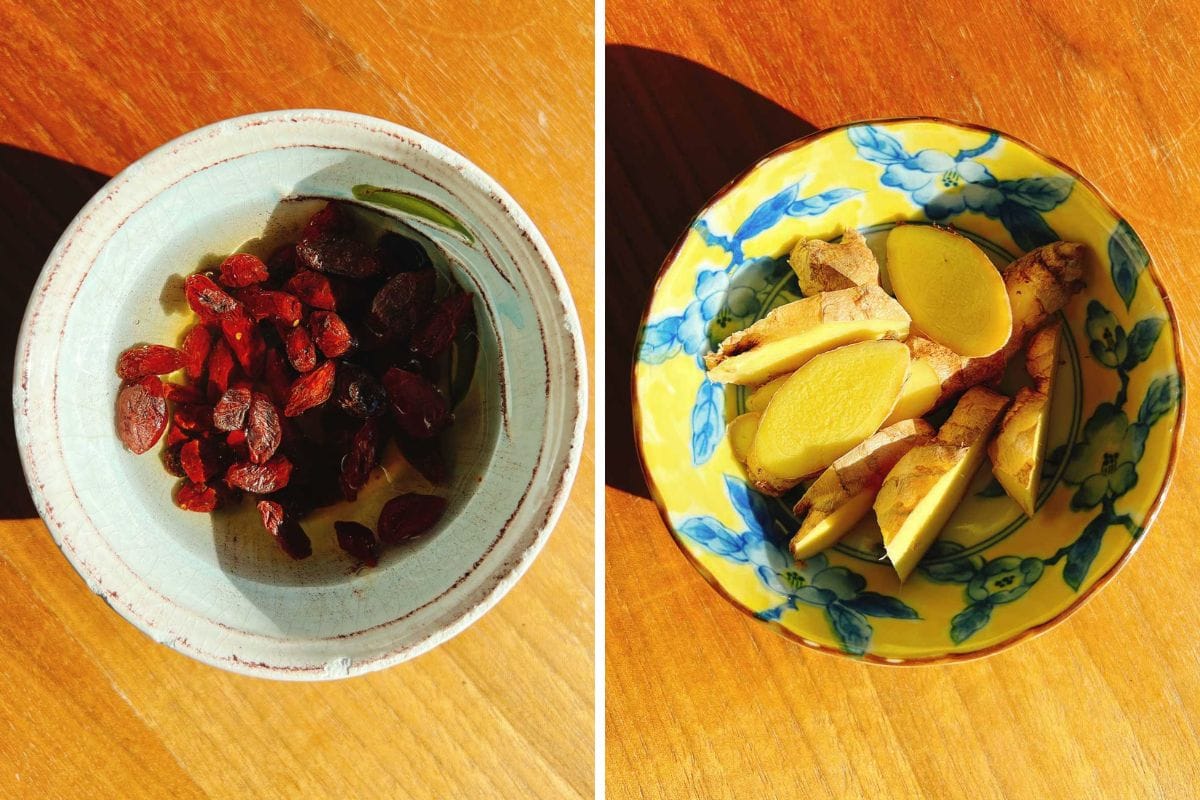 Two ceramic bowls on a wooden surface; the left holds dried red goji berries soaked in rice wine, perfect for adding to sesame oil chicken soup, while the right displays sliced fresh ginger root in a yellow floral-patterned dish.