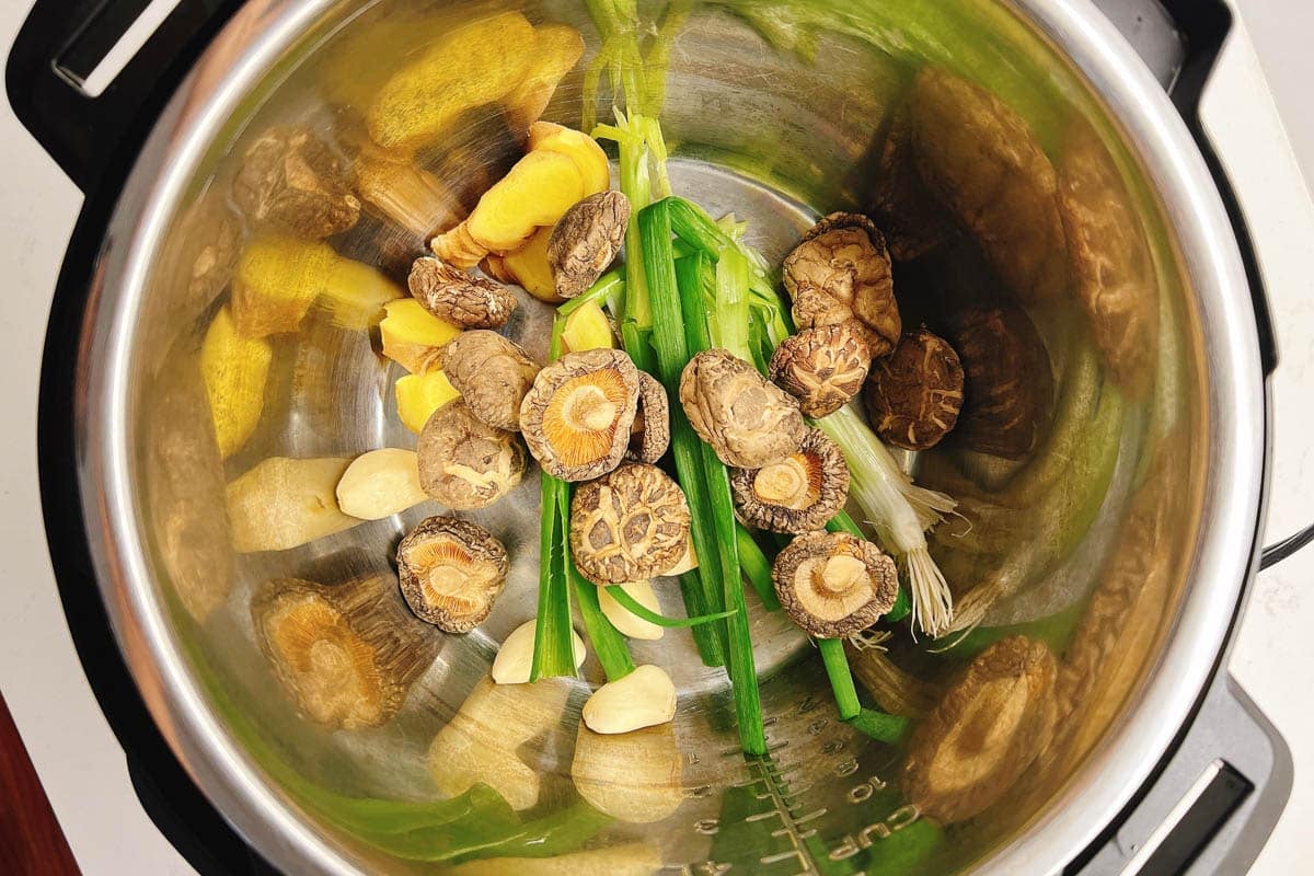 An overhead view of a slow cooker filled with fresh ginger slices, shiitake mushrooms, green onions, and garlic cloves, ready to be cooked for a flavorful Cornish Hen Soup.