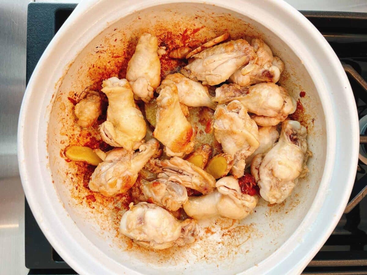Chicken drumettes and wings are cooking in a white clay pot with sliced ginger and black sesame oil, creating a flavorful sesame oil chicken soup. The lightly browned chicken is surrounded by oil and aromatic spices on the stovetop.