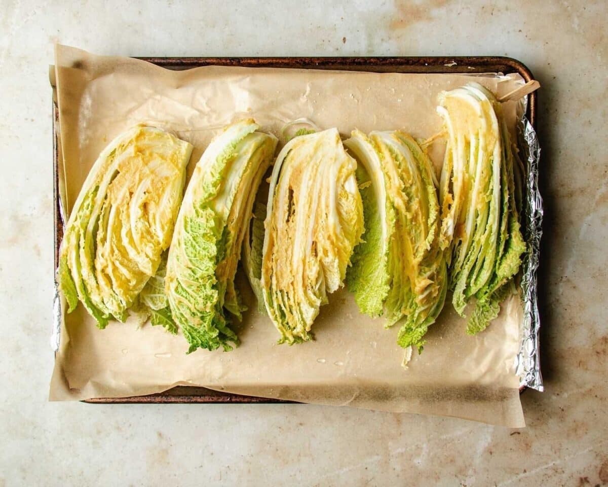 Four wedges of roasted napa cabbage are arranged in a row on a parchment-lined, foil-covered baking sheet. The cabbage has light green and yellow leaves with crisp, layered textures—perfect for roasting.