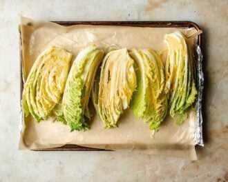 Four wedges of roasted napa cabbage are arranged in a row on a parchment-lined, foil-covered baking sheet. The cabbage has light green and yellow leaves with crisp, layered textures—perfect for roasting.
